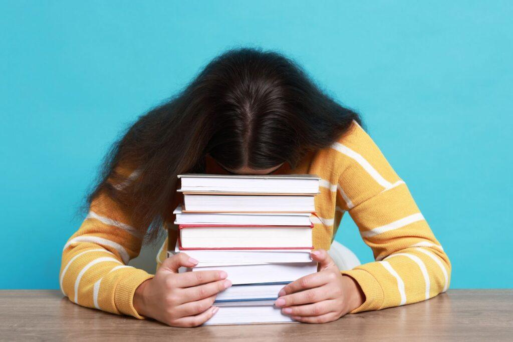 Eine erschöpfte Studentin stuetzt den Kopf auf einen Stapel Buecher, Symbol fuer Schreibstress und Hilfe durch ghostwriter hausarbeit