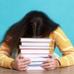 Eine erschöpfte Studentin stuetzt den Kopf auf einen Stapel Buecher, Symbol fuer Schreibstress und Hilfe durch ghostwriter hausarbeit