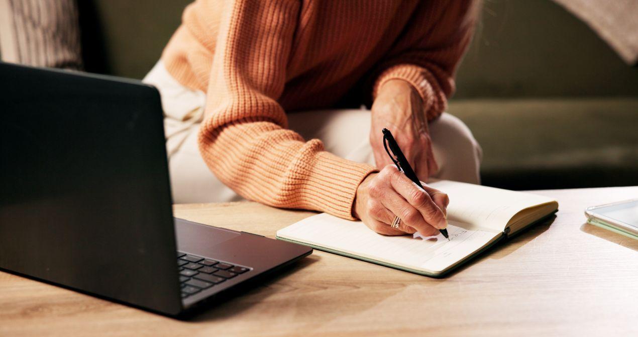 Eine Person schreibt konzentriert in ein Notizbuch neben einem Laptop, steht fuer Struktur und Organisation beim ghostwriter hausarbeit Projekt
